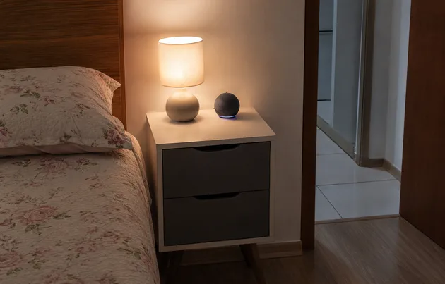 Smart speaker sobre criado-mudo em quarto de apartamento pequeno, ao lado de abajur aceso